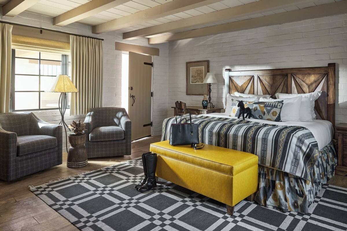 Un dormitorio con alfombra a cuadros, cama con ropa de cama estampada, cabecero de madera, reposapiés amarillo y una lámpara.
