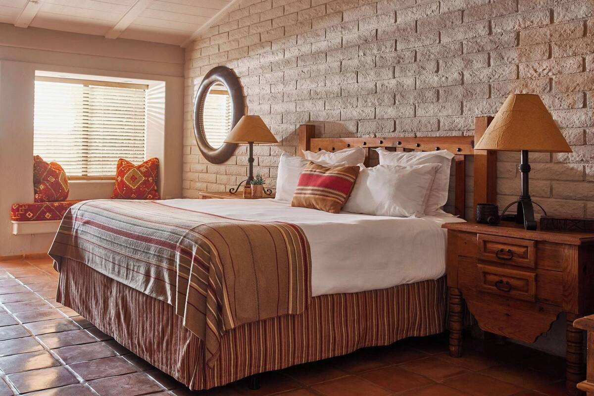 Un dormitorio con pared de ladrillo, un espejo redondo, una cama con ropa de cama a rayas, dos mesitas de noche con lámparas y un banco con cojines.