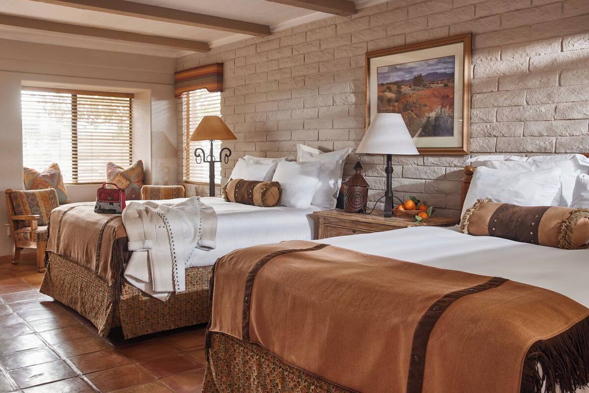 Habitación de hotel con dos camas, un ventanal grande con persianas, un cuadro colgado en la pared y suelo de madera.