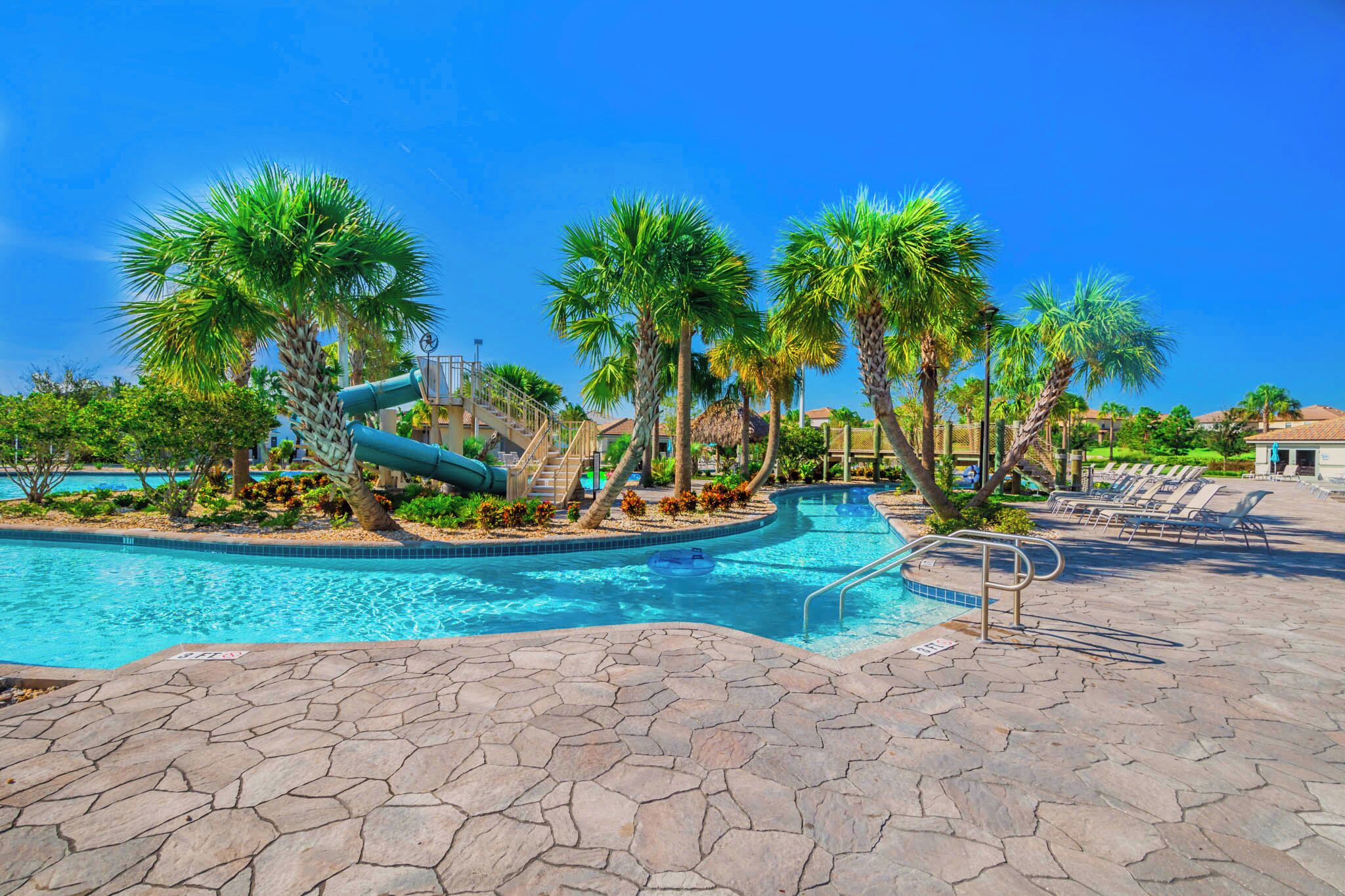 A swimming pool with a slide, palm trees, lounge chairs, and a stone patio.