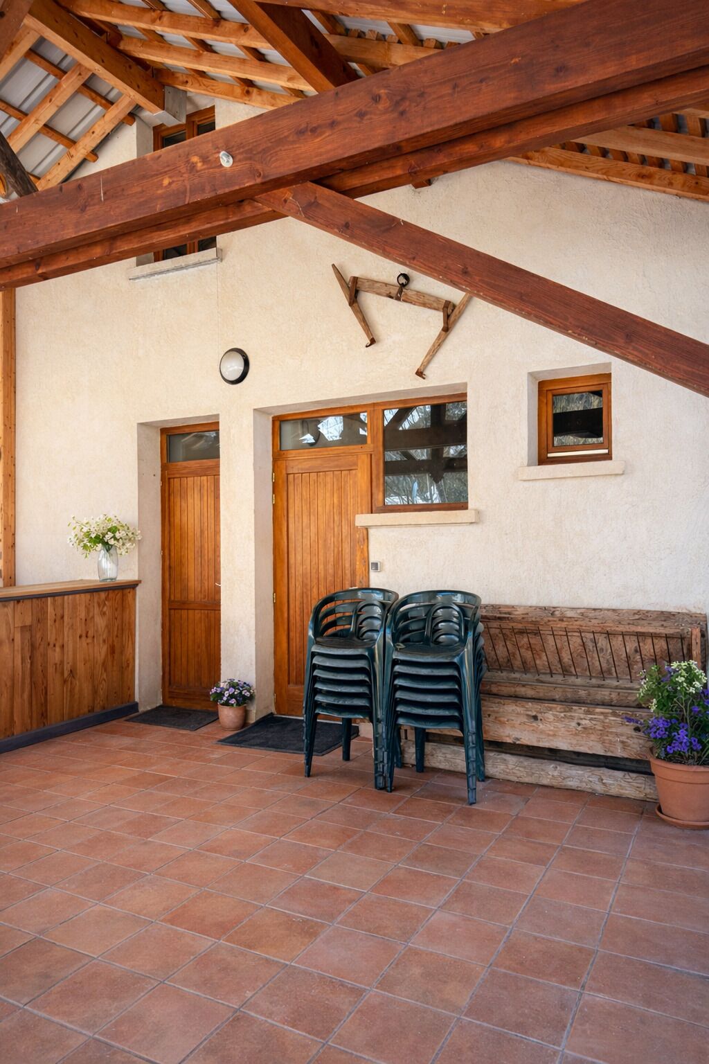 Un espacio al aire libre rústico con un banco de madera, dos sillas de plástico, una puerta de madera y un ventanal pequeño.