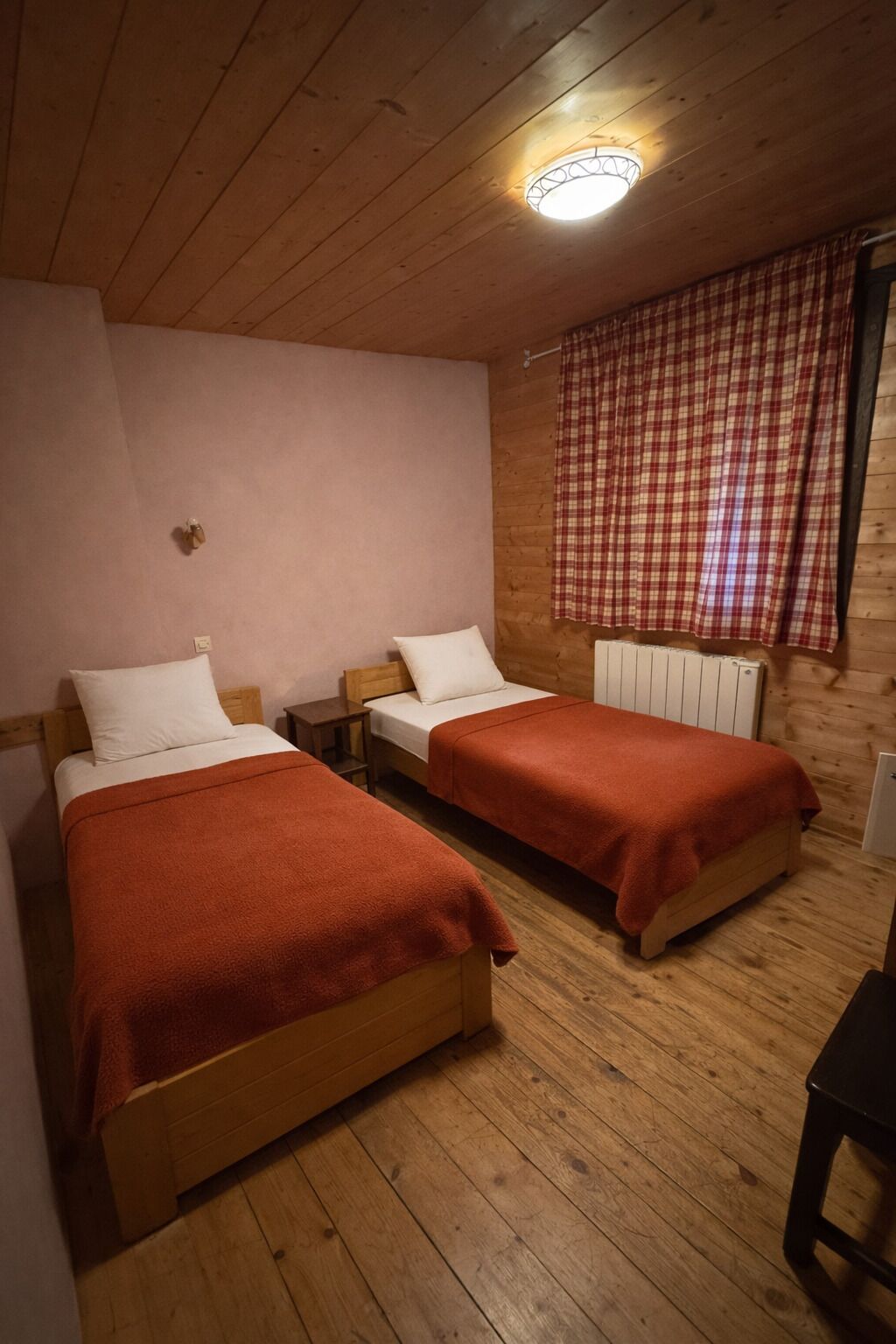 A room with two wooden beds, a radiator, a window with checkered curtains, and a ceiling light fixture.