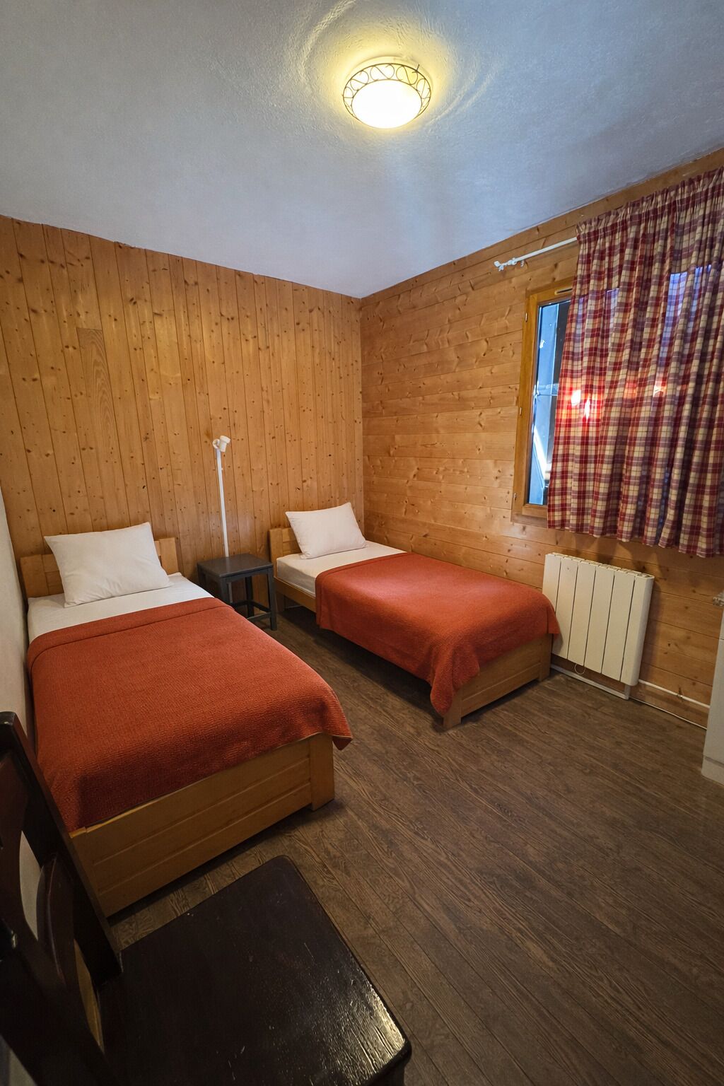 Una habitación con dos camas de madera, una mesita de noche, un radiador y una ventana con cortinas a cuadros.