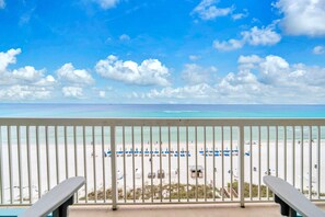 Blick auf einen Strand vom Balkon mit Geländer, strahlend blauer Himmel und ein Sandstrand mit Sonnenschirmen und Liegestühlen.