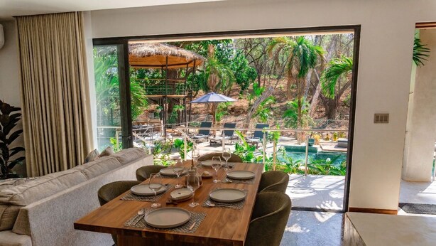 Ein Essbereich mit einem Holztisch, gedeckt für sechs Personen, einer Couch und Blick auf ein Schwimmbad und einen tropischen Garten.