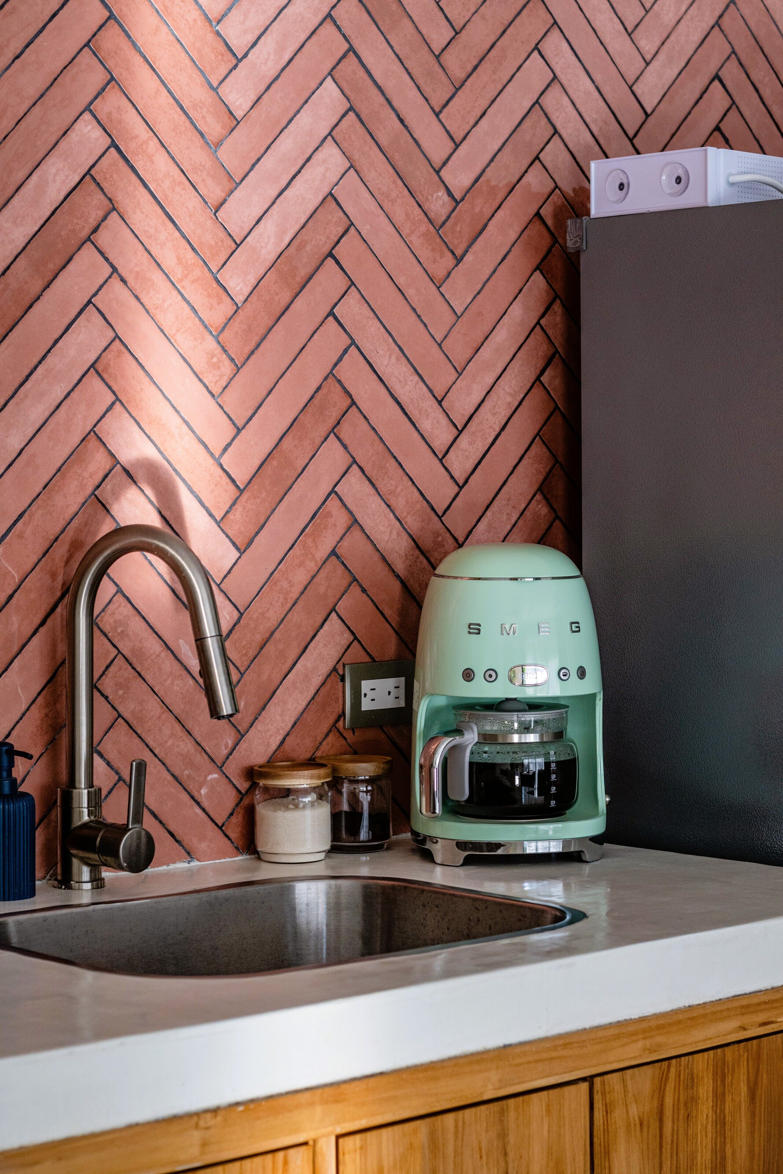 A kitchen with a herringbone-patterned backsplash, a stainless steel sink, a green coffee maker, and wooden cabinets.