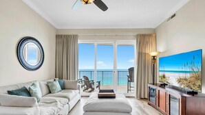 A living room with a sofa, ottoman, and TV, a balcony with chairs, and a large window with a view of the beach. - Ocean Villa Penthouse 2306 (Panama City Beach)