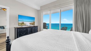 A bedroom with a large bed, a TV, a ceiling fan, and a balcony with a view of the ocean. - Ocean Villa Penthouse 2306 (Panama City Beach)