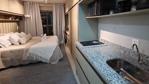 Een compacte hotelkamer met een bed, kitchenette en uitzicht op de stad 's nachts.