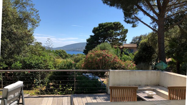 En træterrasse med bænk og bord, med udsigt over en have med træer og hav.