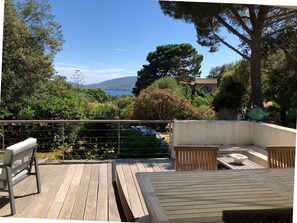 Eine Holzdachterrasse mit einer Bank und einem Tisch, die auf einen Garten mit Bäumen und Meerblick blickt.