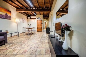 Ein Zimmer mit Steinfußboden, Holzbalken an der Decke, einem Essbereich mit Stühlen und Tisch, einer Couch und einer großen weißen Vase mit Blumen.
