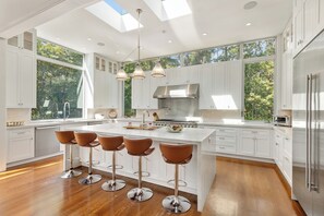 A modern kitchen with a central island, bar stools, and large windows. - Luxury 6-Bedroom Gated Estate on 5 Acres with Pool, Tennis, Gym & Privacy (Bridgehampton)