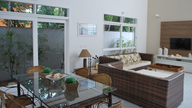 Un salon moderne avec une table à manger en verre, des meubles en osier et un téléviseur à écran plat fixé au mur.
