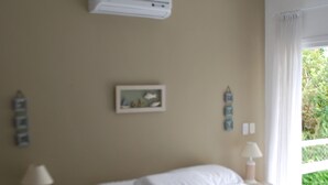 Une chambre à coucher comprenant un lit, des tables de chevet, un climatiseur, une fenêtre avec des rideaux et un tableau accroché au mur.