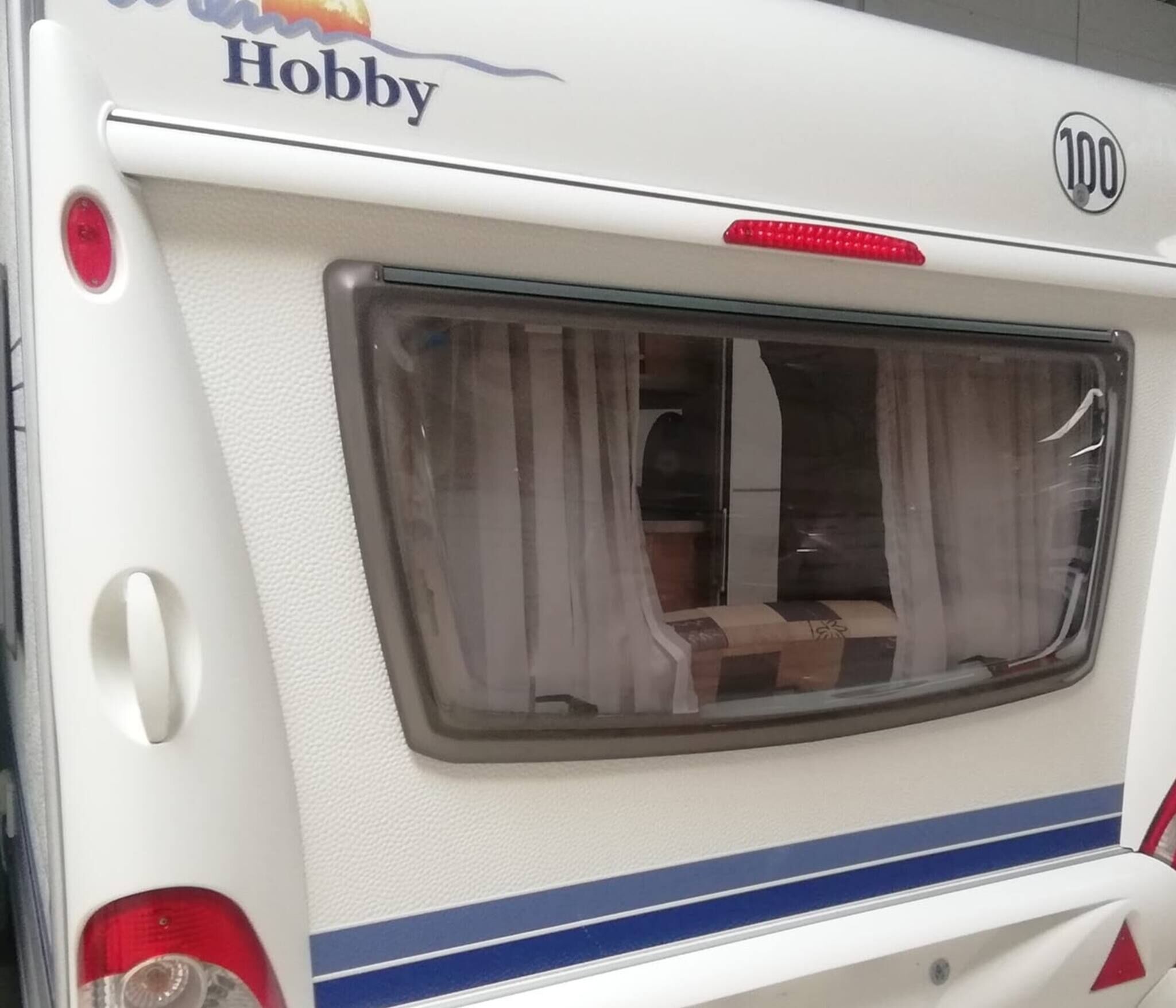 A white RV with a 'Hobby' logo, a rear window with curtains, and a blue stripe.