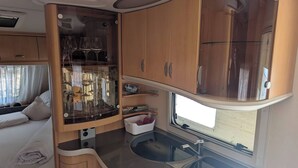 A compact RV kitchen with a built-in sink, a glass cabinet with wine glasses, and a window providing natural light. - Charming Mobile Home Retreat in Ebbs, Austria - Tranquil Escape (Ebbs)