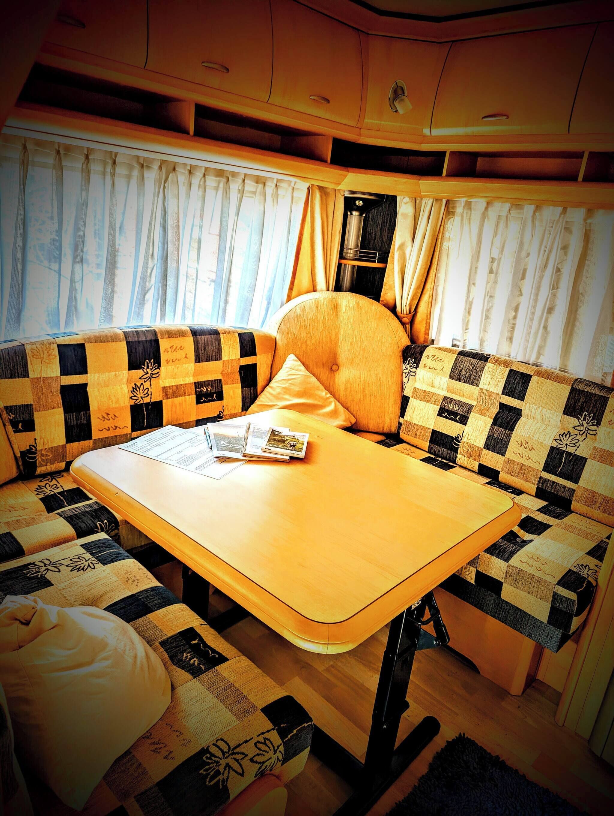 Een gezellig interieur van een camper met een houten tafel, geruit kussens en een raam met gordijnen.