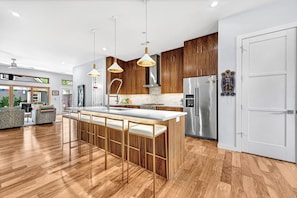 A modern kitchen with wooden cabinets, a stainless steel refrigerator, and a bar area with stools. - Casa La Rosa (Bentonville)