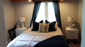 A bedroom with a bed, two bedside tables with lamps, a chair, and a window with curtains. - Charming 2-bedroom Lake Cottage (Durhamville)