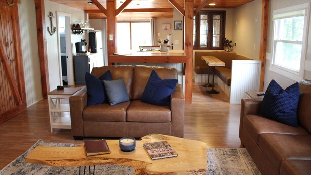 A cozy living room with a brown leather sofa, a wooden coffee table, and a kitchen in the background. - Charming 2-bedroom Lake Cottage (Durhamville)