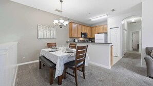 A dining area with a table set for four, a kitchen with wooden cabinets, a microwave, and a refrigerator. - Sunshine Stay Near Disney  2BR  Walk to Pool (Kissimmee)