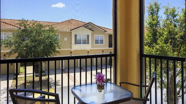 A balcony with a glass table and chairs, a flower vase, and a view of a building with a tiled roof. - Sunshine Stay Near Disney  2BR  Walk to Pool (Kissimmee)