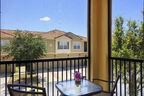 Un balcon avec une table et des chaises en verre, un vase de fleurs et une vue sur un bâtiment doté d’un toit en tuiles.