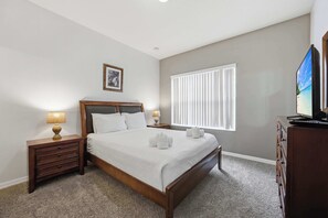 Ein Schlafzimmer mit Bett, Nachttischen, einem Schminktisch, einem Fernseher, einem Fenster mit Jalousien und einem gerahmten Bild an der Wand.