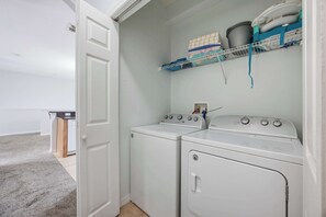 Una sala de bugaderia amb dues rentadora frontals blanques, una estant amb roba doblada i una porta blanca.