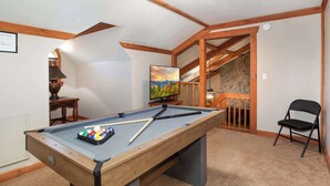 Uma mesa de bilhar com bolas, uma televisão e uma cadeira em um quarto com vigas de madeira e parede de pedra.