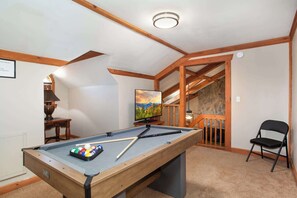 Ein Billardtisch mit Kugeln, ein Fernseher und ein Sessel in einem Raum mit Holzbalken und einer Steinwand.