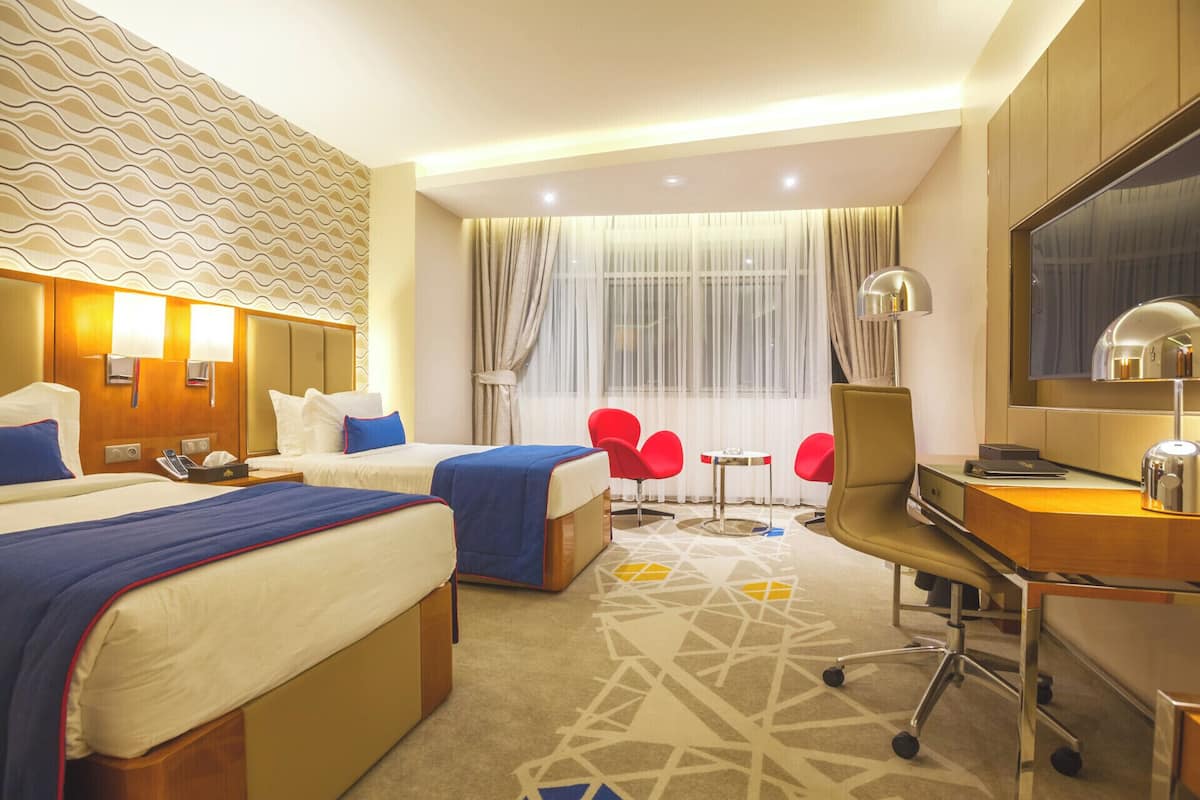 A hotel room with two beds, a desk, a chair, a TV, and a window with curtains.