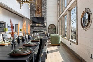 A dining area with a long table set for a meal, a stone fireplace, and large windows. - 7-Bedroom Luxury Cabin with Indoor Pool, Rooftop & Pickleball Court (Sevierville)