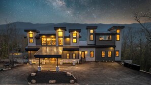 A modern multi-story house with a large driveway, stone foundation, and multiple illuminated windows.
