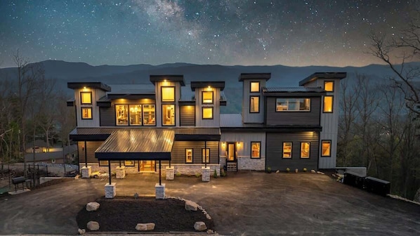 A modern multi-story house with a covered patio and a starry night sky. - 7-Bedroom Luxury Cabin with Indoor Pool, Rooftop & Pickleball Court (Sevierville)