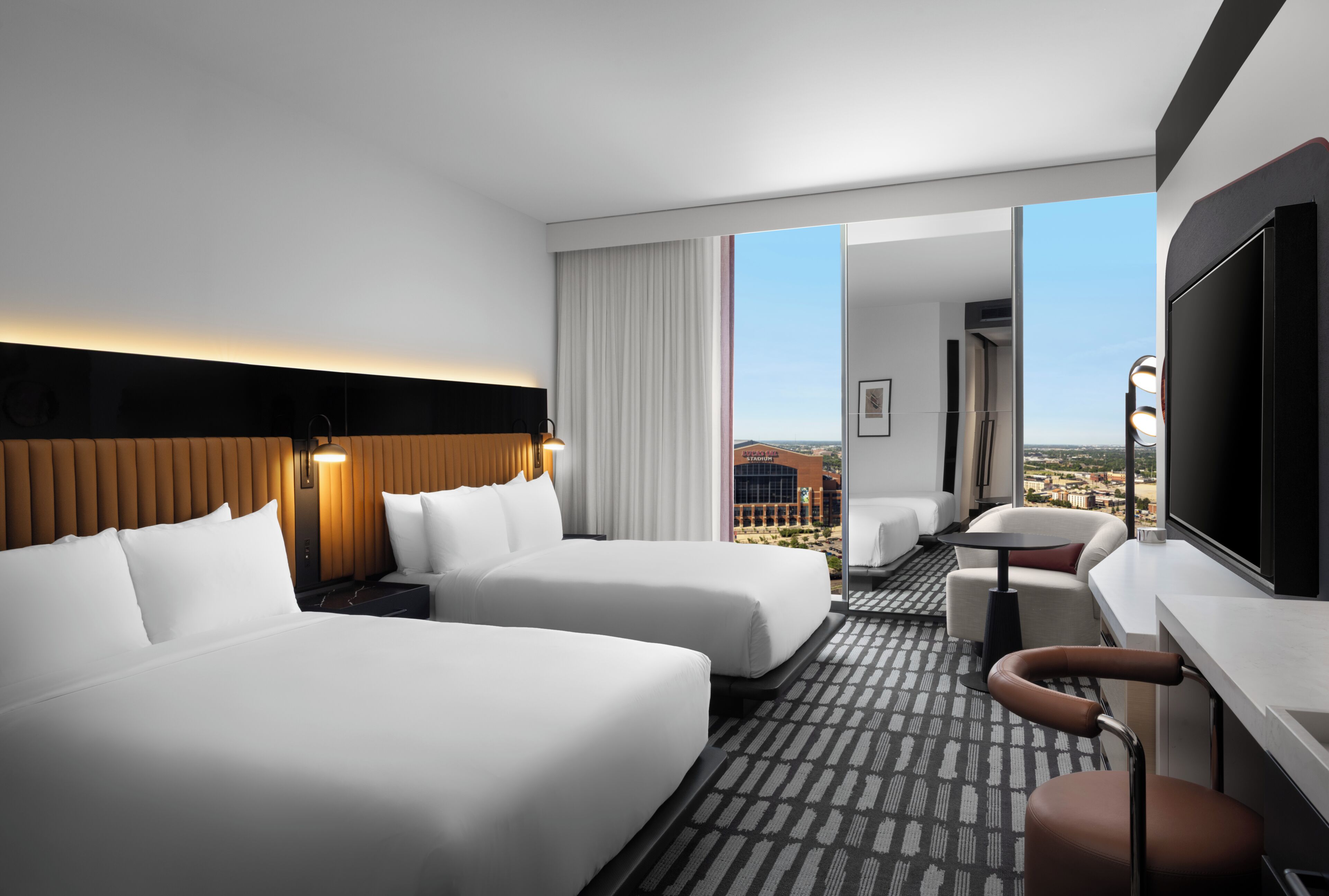 A hotel room with two beds, a desk, a TV, and a view of the city.