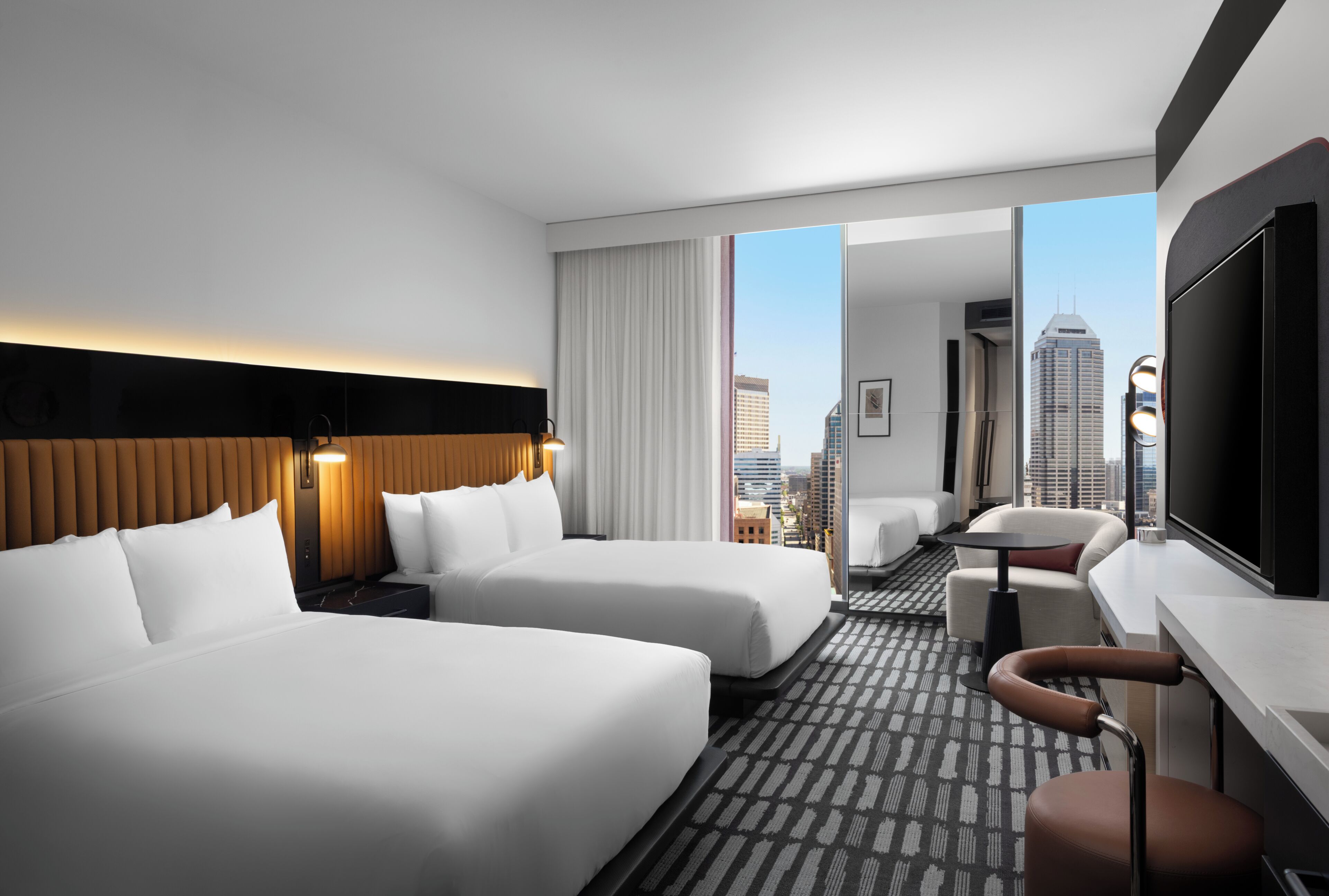 A hotel room with two beds, a desk, a chair, and a large window offering a city view.