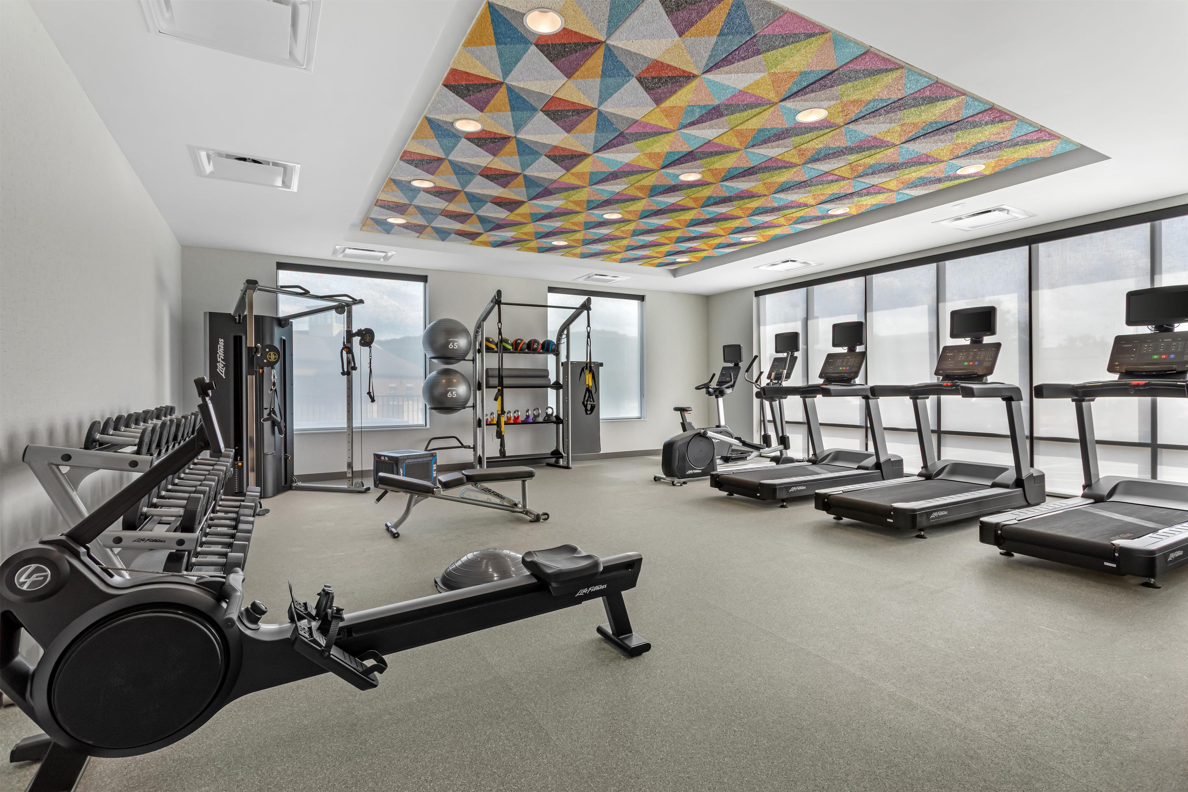 A gym with various exercise equipment, large windows, and a colorful geometric ceiling.