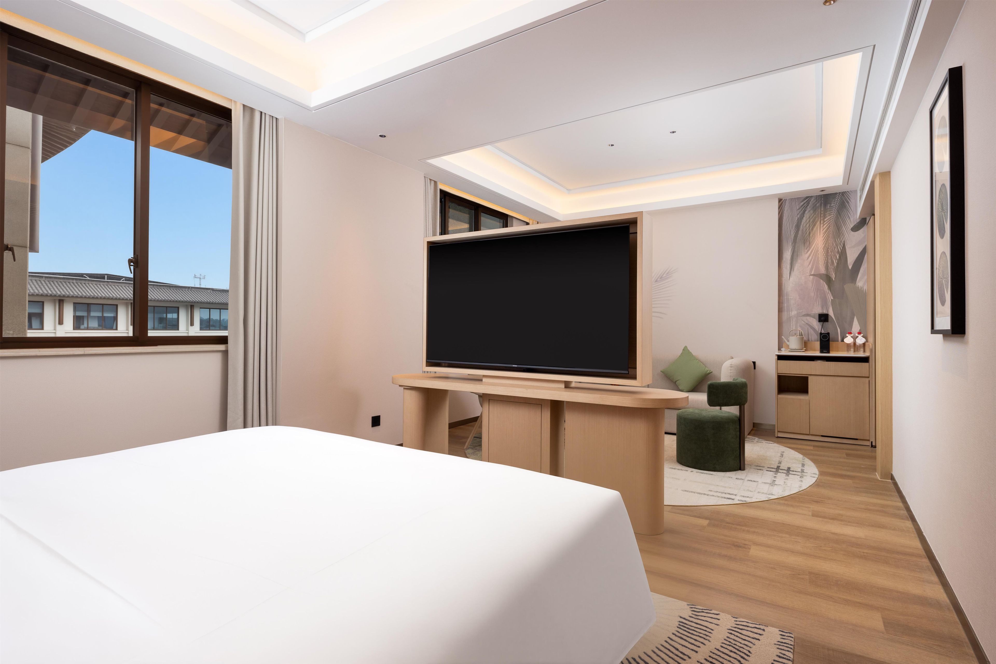 A modern hotel room with a large bed, a flat-screen TV, a small kitchen area, and a view of the outside.