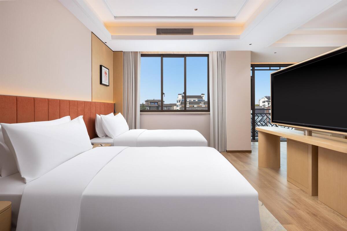 Ein Hotelzimmer mit zwei Betten, einem großen Fenster mit Blick auf die Stadt, einem Flachbildfernseher und einem Holztisch.