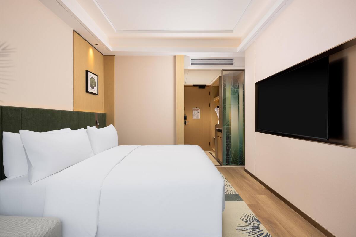 Ein Hotelzimmer mit einem Bett, einem Flachbildfernseher, einem gerahmten Bild an der Wand und einem Flur, der zu einem Badezimmer führt.
