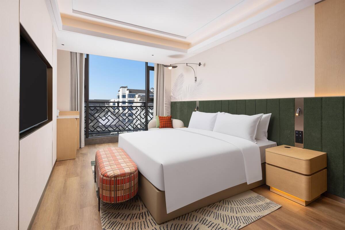 Ein modernes Hotelzimmer mit einem großen Bett, einer kleinen Bank, einem Nachttisch und Blick auf die Stadt.