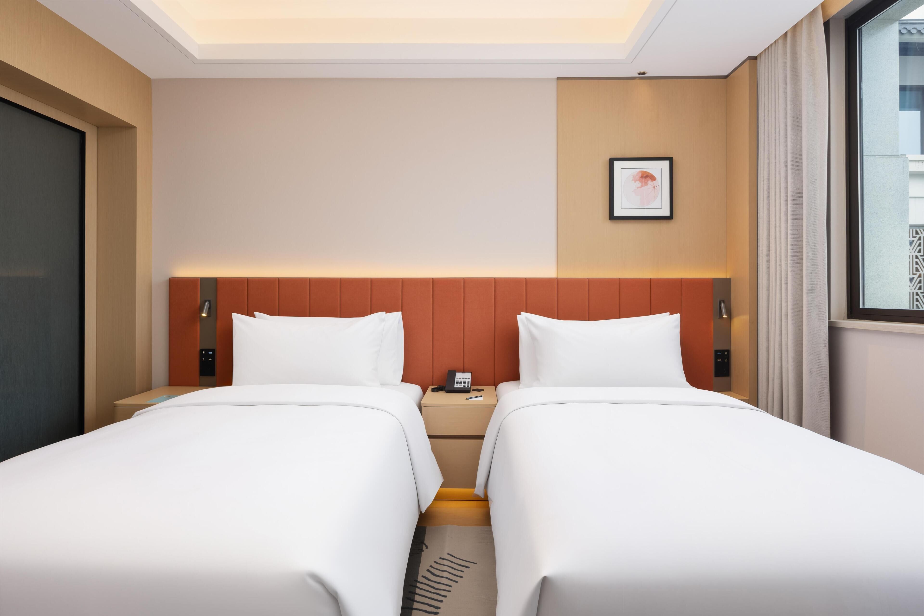 A hotel room with two beds, a nightstand with a phone, a framed picture on the wall, and a window with curtains.