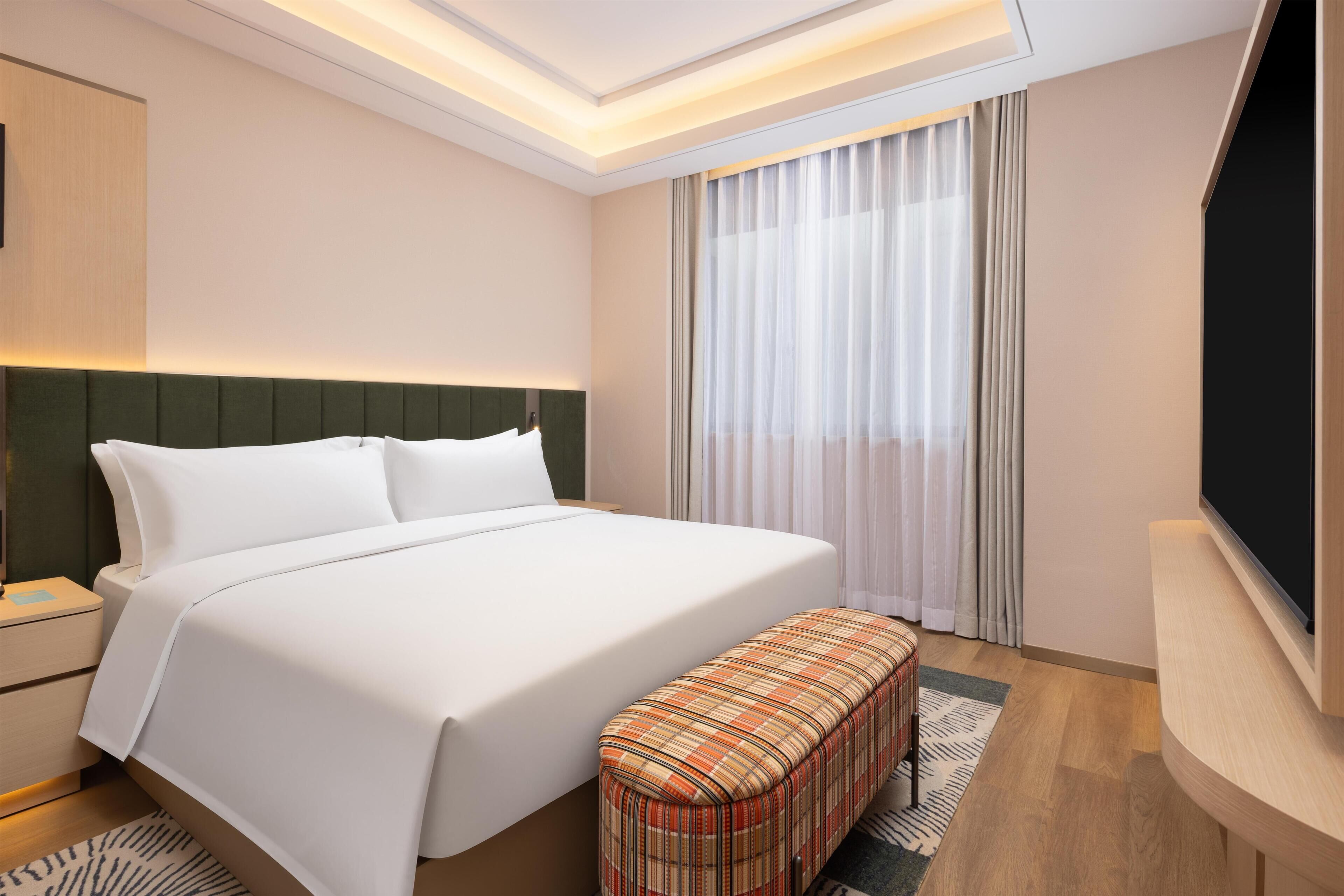 A hotel room with a bed, a bench, a TV, a nightstand, and a window with curtains.