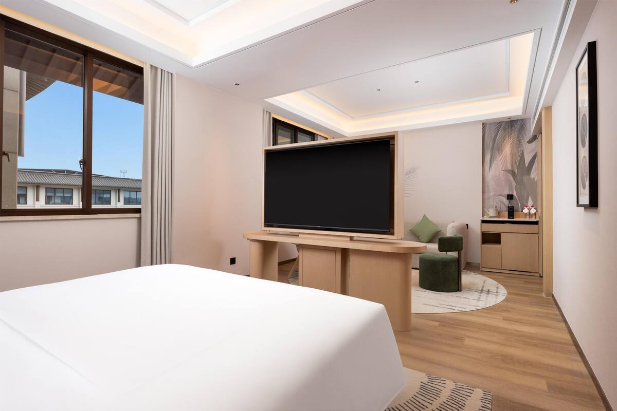 Ein modernes Hotelzimmer mit einem großen Bett, einem Flachbildfernseher, einer kleinen Küchenzeile und Blick auf umliegende Gebäude.