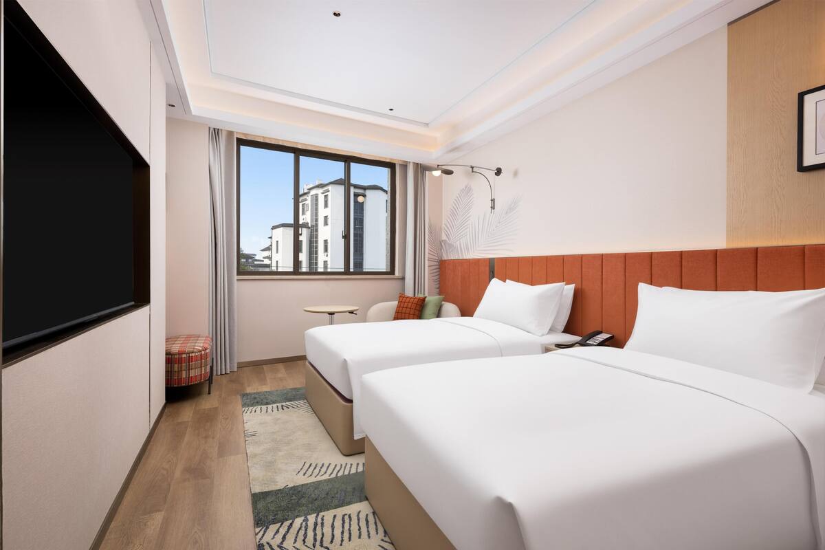 Ein Hotelzimmer mit zwei Betten, einem Fernseher, einem Fenster mit Blick auf Gebäude und einem kleinen Tisch.