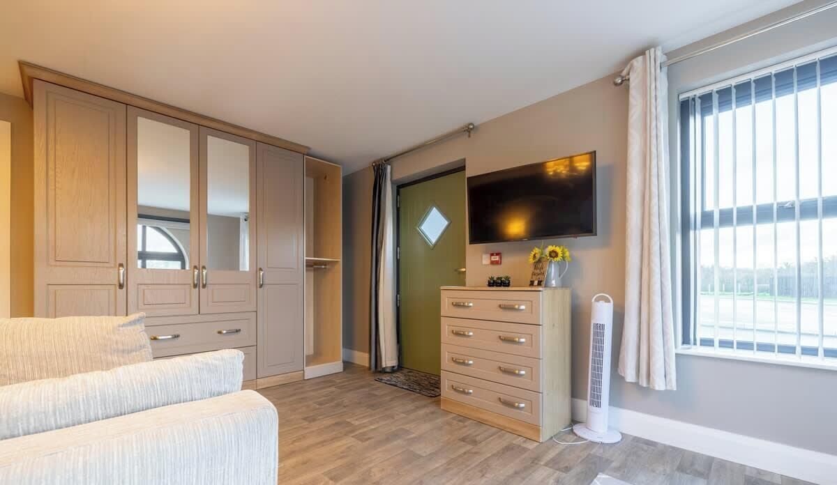 A hotel room with a large wardrobe, a flat-screen TV mounted on the wall, a small dresser, and a window with curtains.