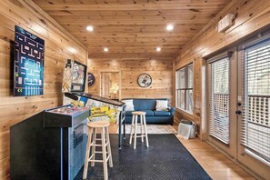 Et værelse med trævægge og -loft, en blå sofa, to træbærestole, et sort gaming-bord og et vægur.