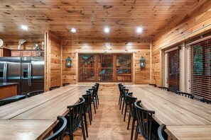 A wooden interior with a long dining table and chairs, a kitchen area with stainless steel appliances, and a large window with blinds. - 7-Bedroom Gatlinburg Cabin with Theater, Two Game Rooms & Hot Tub (Gatlinburg)